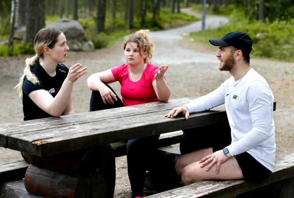 Tre studenter i träningskläder sitter runt ett picknickbord i skogen och pratar