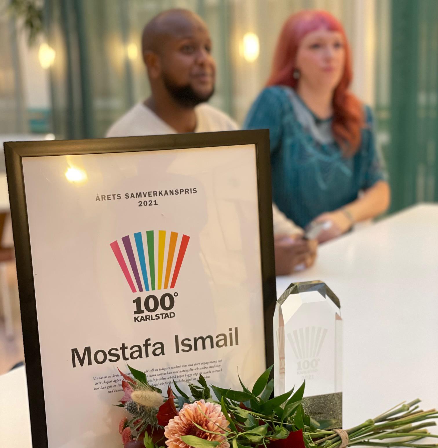 Inramat diplom och en glasstatyett stående på ett bord med blommar framför. Två personer i bakgrunden. 