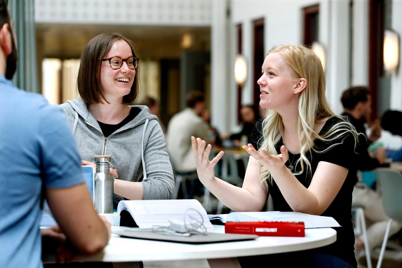 Tre studenter kring fikabordet diskuterar och gestikulerar