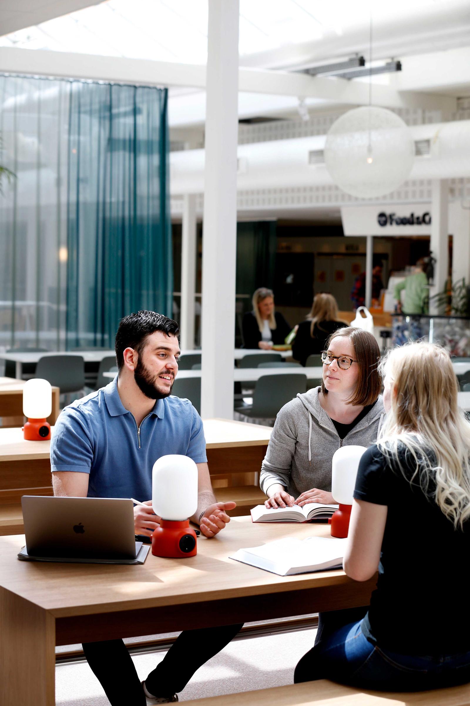 Tre studenter kring fikabordet diskuterar och gestikulerar