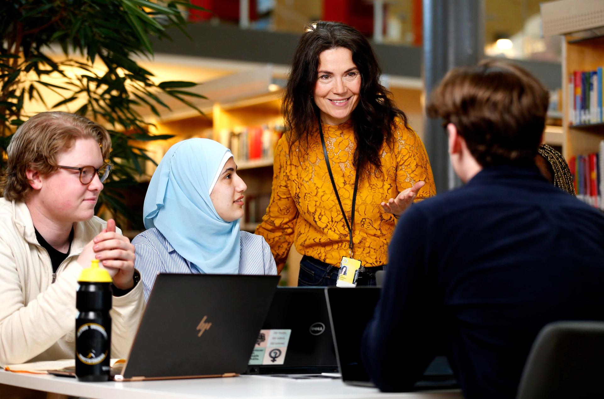 Lärare förklarar och gestikulerar för tre sittande studenter i biblioteket