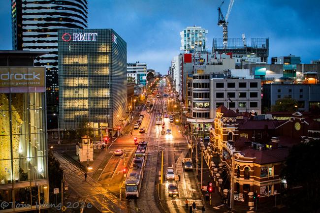 Gatuvy fotograferad ovanifrån en trafikerad gata i Melbourne. Det är natt och vägen sträcker sig mot horisonten. På gatan lyser bilar spårvagnar och trafikljus. På vägens sidor syns belysta höghus och på ett av husen till vänster i bildens förgrund syns en neonskylt med texten RMIT högst upp på husets fasad.