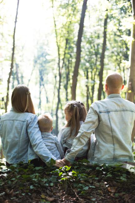 En familj med mamma, pappa och två brn sitter med ryggen mot kameran i en lummig skog