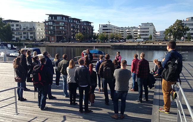 En grupp med tjugotalet deltagare lyssnar på två guider under en riskvandring. Gruppen står på en träbrygga i Karlstads Inre hamn. I bakgrund synd bostadsbebyggelse på andra sidan av hamnbassängen.
