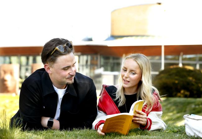 en kille och tjej ligger i parken utanför huvudentrén och läser i en bok.