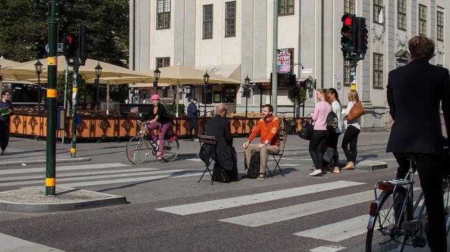 En gata med cyklister, gångtrafikanter och två personer som sitter på stolar mittemot varandra på övergångsstället.