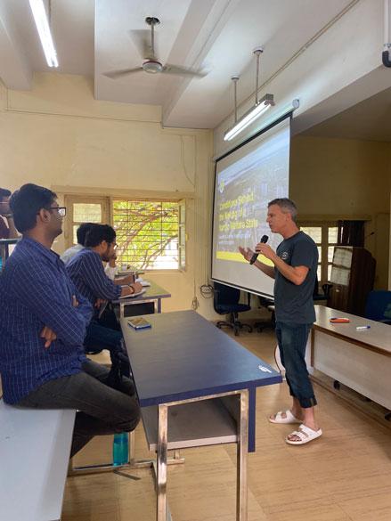 En amn står vid en powerpoint bild och pratar inför åhörare i ett klassrum i Varansai